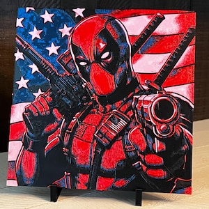 May include: Square canvas art depicting Deadpool in a red and black suit, holding guns, set against an American flag. The artwork is displayed on a black easel, with a patriotic and action theme.