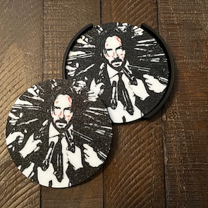 Può includere: Due sottobicchieri rotondi neri con una grafica in bianco e nero di un uomo con pistole. L'uomo ha sangue sul viso. I sottobicchieri sono su una superficie di legno.