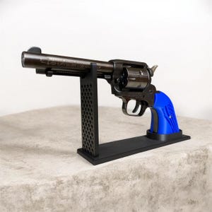 Könnte beinhalten: Ein metallischer Revolver mit dunklem Finish und einem leuchtend blauen Griff mit strukturiertem Muster. Die Schusswaffe ist auf einem schwarzen Präsentationsständer mit vertikaler Gitterstütze und rechteckiger Basis horizontal montiert.