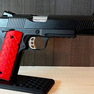 以下が含まれることがあります： 銀色のスライドと赤いテクスチャグリップを備えた黒い拳銃。 銃器は黒いスタンドに展示されています。 「IAS KNOXVILLE TN 37920-23402230」というテキストが銃の側面に表示されています。