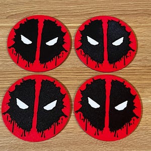 May include: Four round coasters featuring the Deadpool mask design. Each coaster has a red border with a black center split vertically, white eyes, and a dripping effect. The coasters are on a wooden surface.