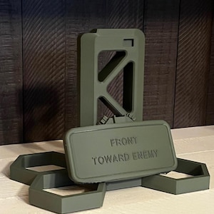 May include: An olive green, rectangular object with the text "FRONT TOWARD ENEMY" is displayed. It is accompanied by a tall, open-frame structure and three hexagonal shapes. The items are arranged on a light-coloured surface.