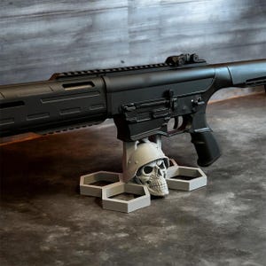 以下が含まれることがあります： 黒いライフルが、軍用ヘルメットをかぶった灰色の頭蓋骨型のスタンドに置かれています。スタンドは六角形のベースです。背景は濃い灰色の表面です。
