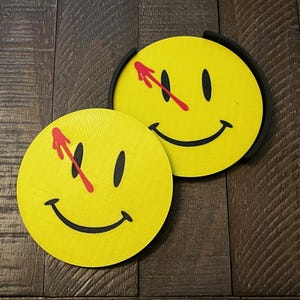 May include: Two yellow smiley face coasters with black eyes and smiles, and a red arrow through the face. One coaster is in a black holder. The coasters are on a dark brown wooden surface.