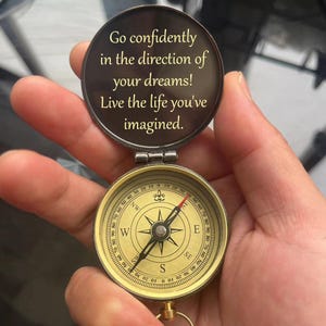 May include: A gold compass with a black needle pointing north. The compass is open and the lid has the inscription "Go confidently in the direction of your dreams! Live the life you've imagined."