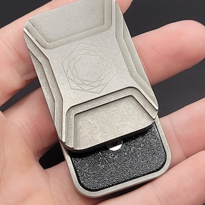 Könnte beinhalten: Ein rechteckiges, metallisch graues Zappelspielzeug mit einem geometrischen Design auf der Oberseite. Der obere Teil lässt sich öffnen und gibt ein strukturiertes schwarzes Innere frei. Das Spielzeug wird in einer Hand gehalten, um seine Größe und sein Design zu zeigen.