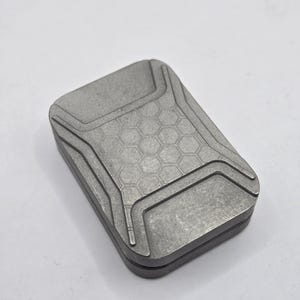 May include: A rectangular, metallic gray fidget slider with a matte finish. The top surface features a central hexagonal grid pattern surrounded by raised, angular border lines. The object consists of two stacked, aligned metal plates.