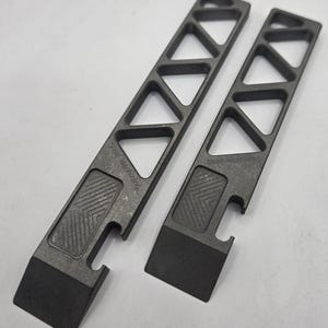 May include: Two dark grey metal pry bars featuring a skeletonised handle with triangular cutouts, a textured grip section, and a small notch near the base. The text "Shed Machining" is engraved on the surface of each tool, which has a flat, angled tip.