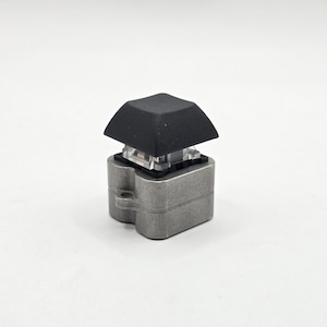 May include: A single, mechanical keyboard keycap. The keycap is black and square, sitting atop a clear, rectangular switch. The base is a silver-colored metal with a unique, curved design. The keycap is isolated against a white background.