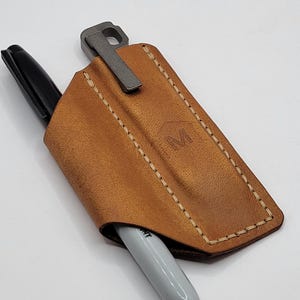May include: A brown leather pen holder with a metal clip, holding a black Sharpie pen and a silver pen. The leather case has a stitched border and a debossed logo. The text at the bottom reads "Prybar and pen not included."