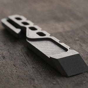 以下が含まれることがあります： マットグレーの金属製マルチツール。面取りされたエッジ、斜めのテクスチャが施されたグリップ部分、背面に沿った一連の長方形の切り抜きが特徴です。平らなベースと先細りのノミのような先端を持つ、幾何学的で角張ったプロファイルです。