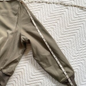 May include: A tan bomber jacket with a ribbed hem and cuffs. A measuring tape is laid across the sleeve, showing a length of approximately 27 inches. The jacket is on a white, patterned surface.