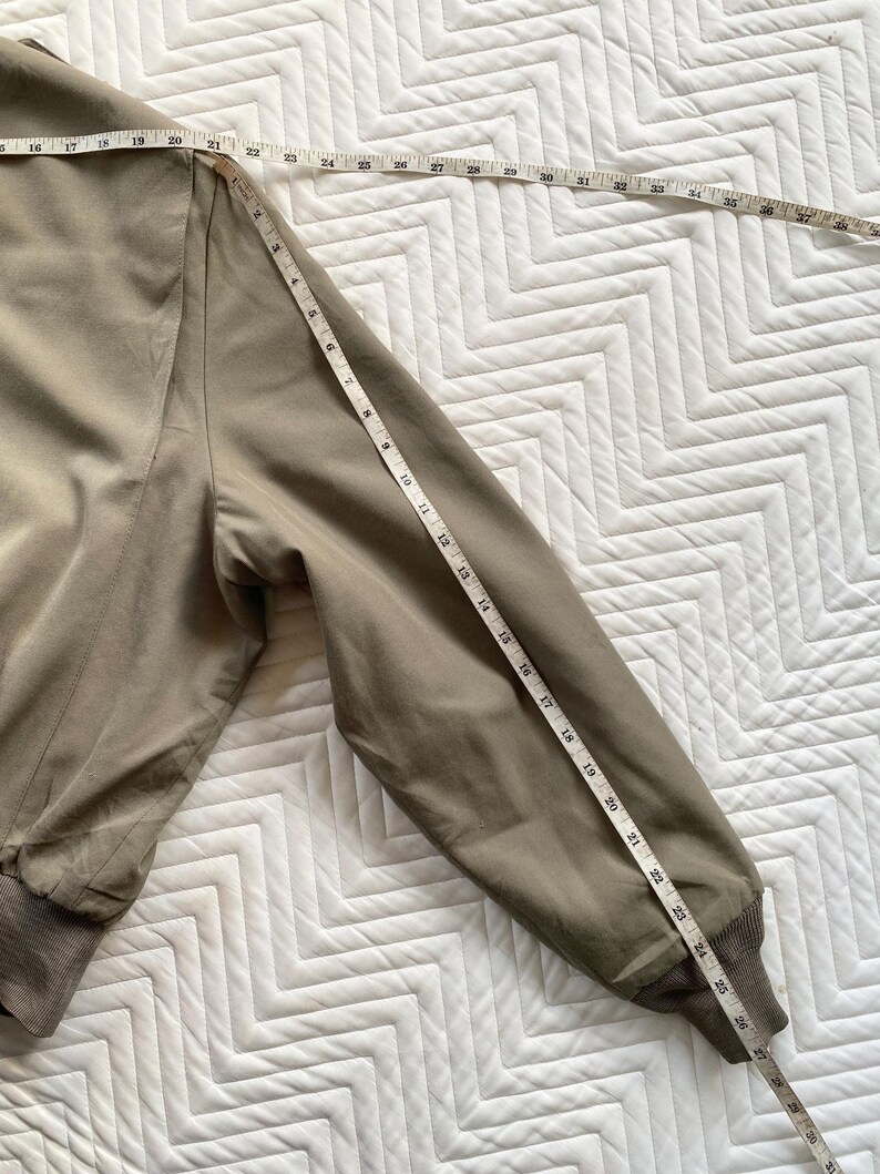 May include: A tan bomber jacket with a ribbed hem and cuffs. A measuring tape is laid across the sleeve, showing a length of approximately 27 inches. The jacket is on a white, patterned surface.