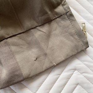 May include: Close-up of a section of khaki-colored fabric, possibly from a garment. The fabric has a textured appearance with visible stitching and a gold-colored zipper. The image shows the edge of the fabric with a small tear.
