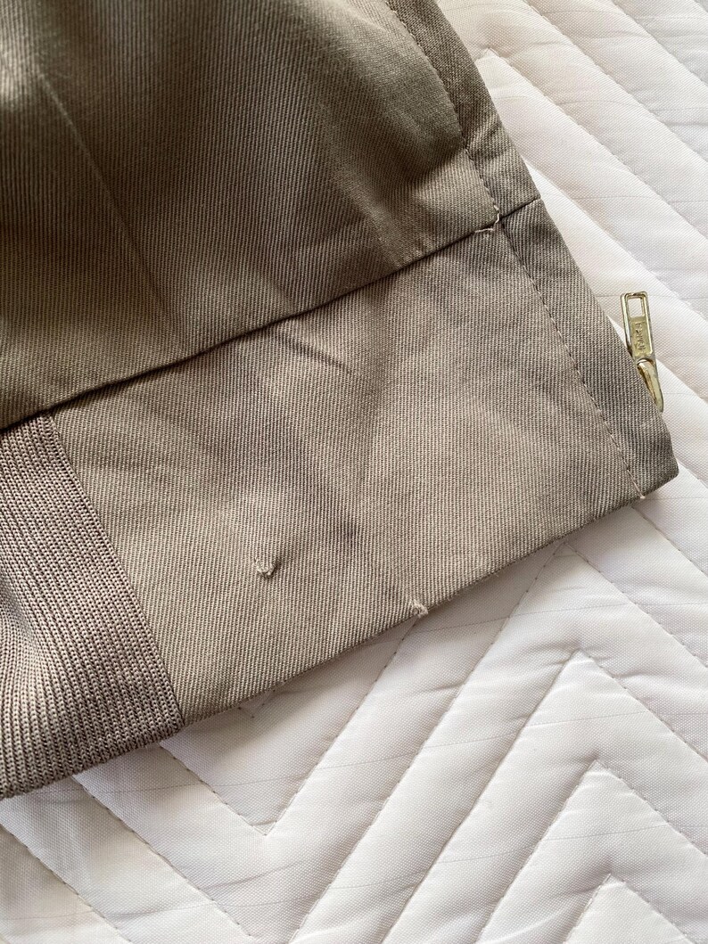 May include: Close-up of a section of khaki-colored fabric, possibly from a garment. The fabric has a textured appearance with visible stitching and a gold-colored zipper. The image shows the edge of the fabric with a small tear.