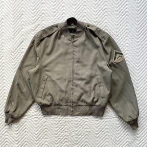 May include: A khaki-colored bomber jacket with a zipper closure, two front pockets, and shoulder epaulets. The jacket features a patch on the left sleeve. The jacket has a ribbed hem and cuffs.