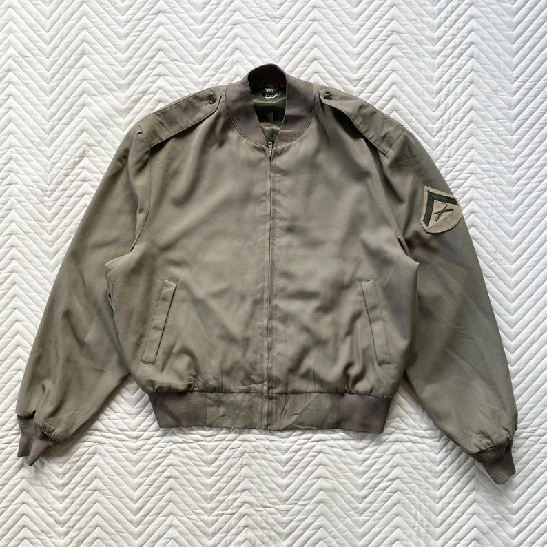 May include: A khaki-colored bomber jacket with a zipper closure, two front pockets, and shoulder epaulets. The jacket features a patch on the left sleeve. The jacket has a ribbed hem and cuffs.