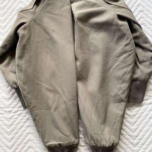 May include: A khaki-colored bomber jacket with a ribbed cuff at the wrist and hem. The jacket has a collar and a smooth, slightly shiny texture. The jacket is laid flat on a white, textured surface.