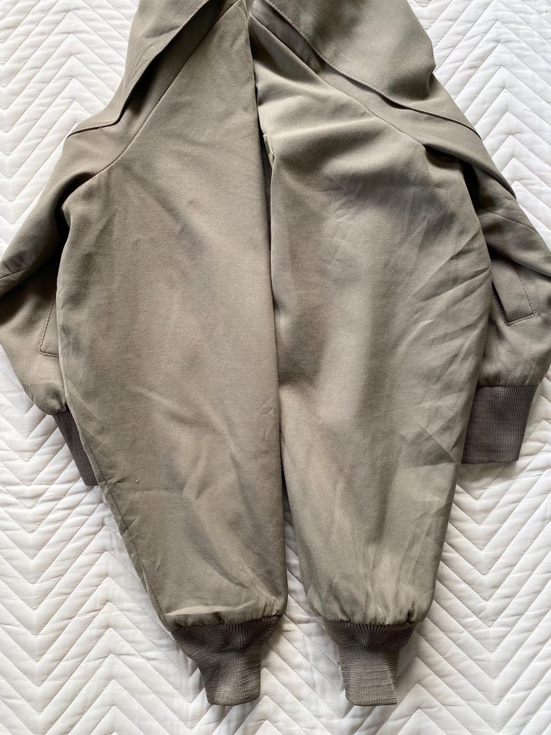 May include: A khaki-colored bomber jacket with a ribbed cuff at the wrist and hem. The jacket has a collar and a smooth, slightly shiny texture. The jacket is laid flat on a white, textured surface.