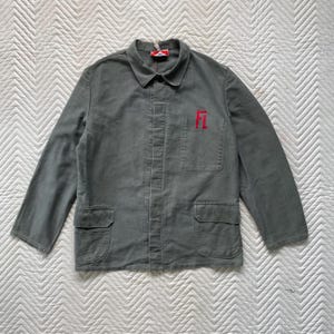 May include: A gray work jacket with long sleeves, a collar, and two front pockets. The jacket has the red letters "FL" embroidered on the chest. The jacket is buttoned up and appears to be made of a durable fabric.