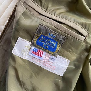 May include: Close-up of the interior of an olive green jacket. The jacket features a label from "Neptune Garment Co. of Boston" and a smaller label with the American flag and text "Made in USA". The jacket also has a pocket.