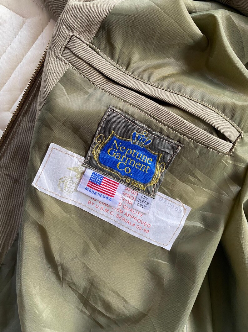 May include: Close-up of the interior of an olive green jacket. The jacket features a label from "Neptune Garment Co. of Boston" and a smaller label with the American flag and text "Made in USA". The jacket also has a pocket.