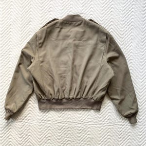 May include: A tan bomber jacket with a ribbed hem and cuffs. The jacket has a stand-up collar and shoulder epaulets. The back of the jacket is visible, showing the seams and the overall shape. The jacket is made of a smooth, possibly synthetic material.