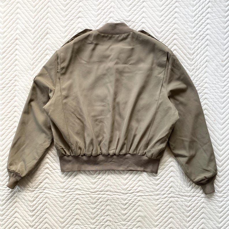 May include: A tan bomber jacket with a ribbed hem and cuffs. The jacket has a stand-up collar and shoulder epaulets. The back of the jacket is visible, showing the seams and the overall shape. The jacket is made of a smooth, possibly synthetic material.