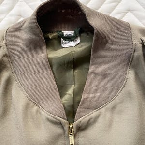 May include: Close-up of a tan bomber jacket with a ribbed collar and cuffs. The jacket has a gold zipper and a green lining. The tag inside the collar reads "Regular".