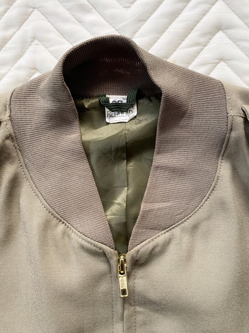 May include: Close-up of a tan bomber jacket with a ribbed collar and cuffs. The jacket has a gold zipper and a green lining. The tag inside the collar reads "Regular".