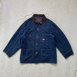 Könnte beinhalten: Blaue Jeansjacke mit Cordkragen. Die Jacke hat vier Vordertaschen, Knopfverschlüsse und ein kleines Etikett auf einer Tasche. Die Jacke liegt flach auf einer weißen Oberfläche.