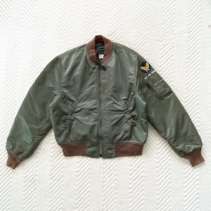 May include: Olive green bomber jacket with brown ribbed cuffs, collar, and waistband. Features a zipper closure, two front pockets, and a U.S. Air Force patch on the left sleeve. The jacket has a classic, military-inspired design.