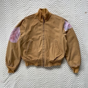 May include: A tan bomber jacket with a high collar and a full zipper closure. The jacket has ribbed cuffs and hem, with a patch of pink and purple fabric on each shoulder. The jacket is made of a durable material.