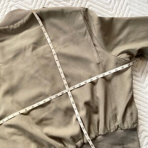 May include: A tan bomber jacket with a ribbed collar and cuffs. A measuring tape is laid across the jacket, showing measurements in inches. The jacket is made of a smooth, lightweight material.