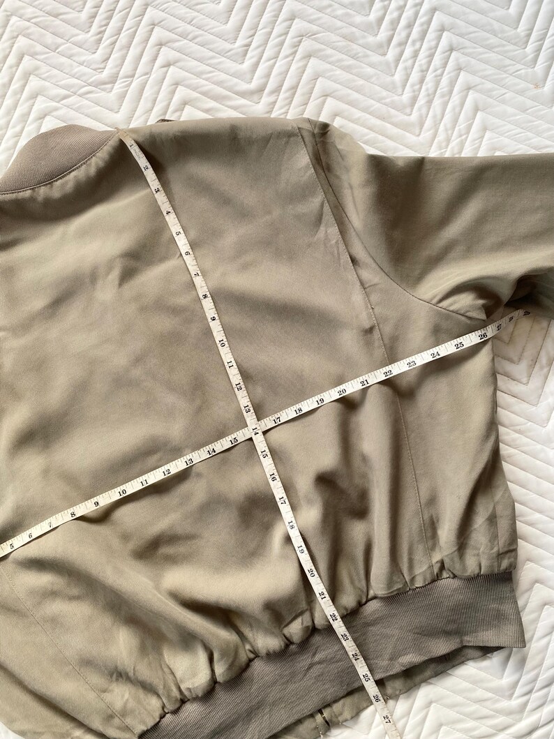 May include: A tan bomber jacket with a ribbed collar and cuffs. A measuring tape is laid across the jacket, showing measurements in inches. The jacket is made of a smooth, lightweight material.