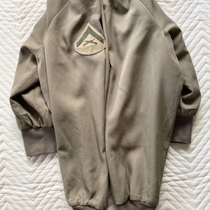 May include: Olive green bomber jacket with a patch on the upper left sleeve. The jacket has ribbed cuffs and hem. The patch features a green chevron design on a tan background. The jacket is made of a smooth, matte fabric.