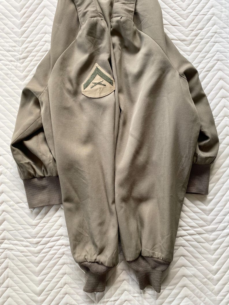May include: Olive green bomber jacket with a patch on the upper left sleeve. The jacket has ribbed cuffs and hem. The patch features a green chevron design on a tan background. The jacket is made of a smooth, matte fabric.