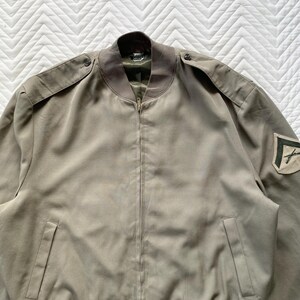 May include: A light olive green bomber jacket with a zip-up front, shoulder epaulets, and a patch on the sleeve. The jacket has a ribbed collar and cuffs, and two front pockets. The jacket is made of a smooth, lightweight fabric.