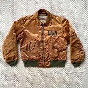 May include: A vintage bomber jacket in a golden-brown colour. It has a zip-up front, two front pockets, and a collar. The jacket features a patch that reads "NAVAL OFFICER". The cuffs and waistband are dark green.