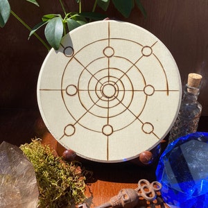 May include: A white wooden circle with a geometric design burned into the surface. The design features a central circle with concentric circles radiating outwards, connected by lines. There are also eight smaller circles evenly spaced around the outer edge of the circle.
