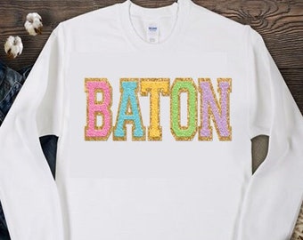 May include: White sweatshirt with the word "BATON" in colorful glitter letters.