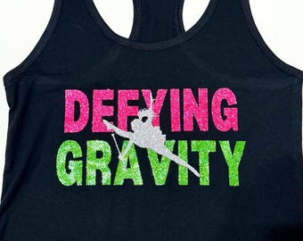 May include: Black tank top with the words "DEFYING GRAVITY" in pink and green glitter. A silhouette of a dancer is in the middle of the words.