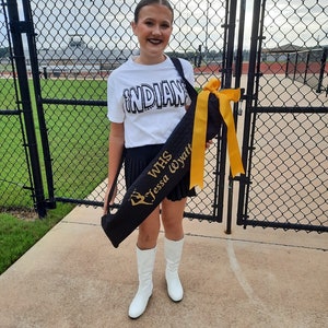 May include: A young person wearing a white shirt with the word "INDIANS" printed on it, black pants, and white boots. They are holding a black bag with a gold sash that says "WHS Tessa Wyatt".