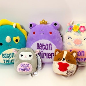 May include: A collection of plush toys in various animal shapes, including a dinosaur, frog, sheep, dog, and pig. The toys are in shades of blue, purple, gray, brown, and white, with embroidered details and the text "Baton Twirler".