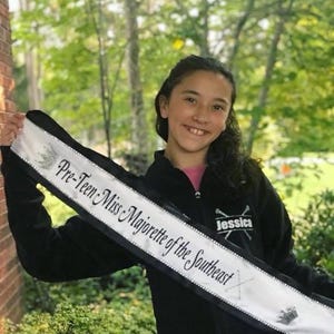 May include: A young person wearing a black jacket with a white sash that reads "Pre-Teen Miss Majorette of the Southeast" and the name "Jessica" embroidered on the jacket.