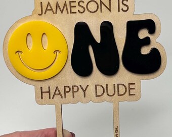 May include: Wooden cake topper with the text "JAMESON IS ONE HAPPY DUDE" and a yellow smiley face. The word "ONE" is in black. The topper has two wooden stakes for insertion into a cake.