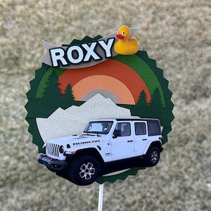 May include: A white Jeep Rubicon cake topper featuring a green, orange and brown mountain scene and a yellow rubber duck. The topper reads "ROXY" in black lettering.