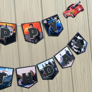 May include: A black and white banner with the word "Happy" spelled out in black letters. Each letter is on a separate pennant with a different image from the Fast and Furious movie franchise. The pennants are strung together with white string.