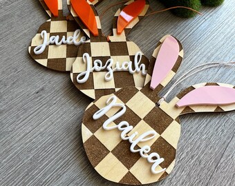 May include: Three wooden bunny-shaped ornaments with checkered patterns and personalized names: "Jada", "Josiah", and "Nailea". The bunny ears are made of orange and pink acrylic.