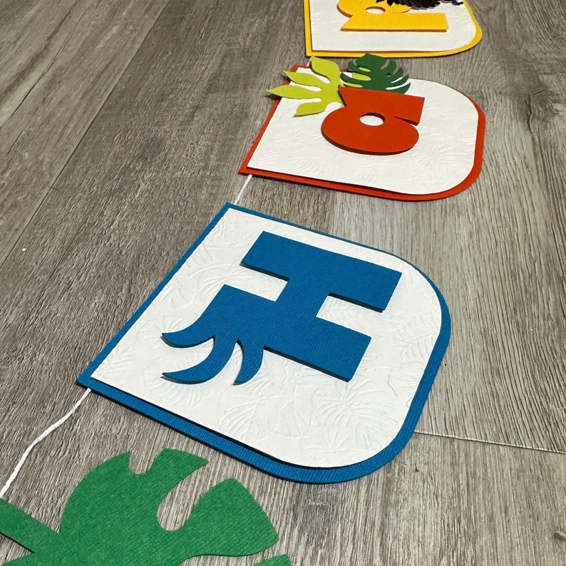May include: A white banner with blue, red, and yellow letters that spell out "Happy" with a green tropical leaf design.